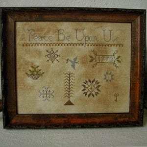 May include: A framed cross-stitch sampler on mottled tan fabric featuring the text Peace Be Upon U, a bird, a tree, geometric star motifs, and the year 1773. The piece is enclosed in a dark, weathered wooden frame with a rustic, distressed finish.