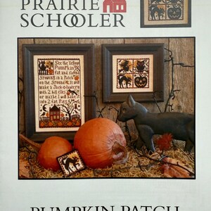 Könnte beinhalten: Buchcover "The Prairie Schooler" mit dem Text "Pumpkin Patch". Das Bild zeigt gerahmte Kreuzstich-Sampler mit Halloween-Themen, Kürbissen und einer schwarzen Katzenfigur. Die Sampler zeigen Bilder eines Spukhauses, Katzen und Kürbissen.