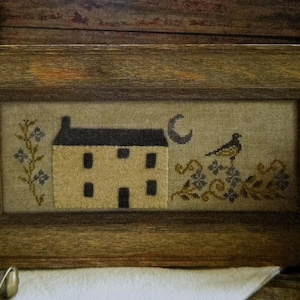 May include: A rectangular framed cross-stitch embroidery featuring a cream-colored house with a dark roof, a crescent moon, a small bird perched on floral vines, and a vertical floral stem, all set against a neutral fabric background.