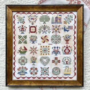 May include: Framed cross-stitch sampler with a gold-colored frame. The sampler features a grid of colorful embroidered motifs, including flowers, hearts, and geometric patterns. The year 1842 is stitched into the design. The border is a scalloped red pattern.