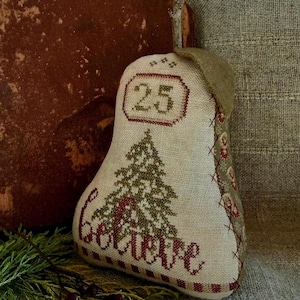 May include: A pear-shaped fabric ornament made of cream-colored linen featuring cross-stitched embroidery. The design includes a green pine tree, the word "believe" in red, and the number "25" in a box. It has a brown felt leaf and a twig stem.