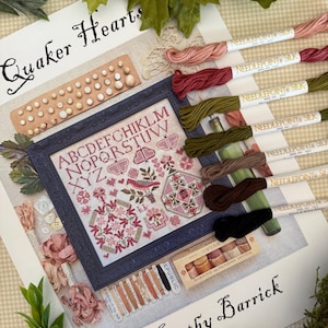 May include: A cross-stitch pattern titled "Quaker Hearts" by Kathy Barrick, featuring a framed sampler with floral and geometric designs in red and pink. Includes various colored threads, buttons, and a box of darning cotton. The image is surrounded by green leaves.