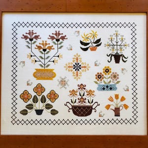 May include: A framed cross-stitch embroidery featuring a variety of colorful flowers in pots. The flowers are stylized and have a vintage aesthetic. The embroidery is surrounded by a black and white border.