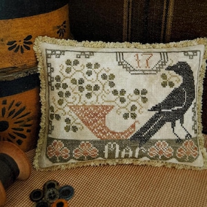 May include: A rectangular fabric pillow with a cross-stitch design featuring a black crow, a cornucopia, and green clover motifs. The embroidery includes the number 17 and the text Mar. The pillow is finished with a beige fringe trim on all edges.