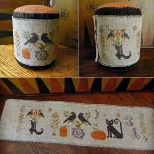 May include: A cylindrical pin cushion with a light beige linen cover featuring cross-stitched motifs of black birds, a black cat, orange pumpkins, and floral elements. The top and bottom edges are finished with a dark brown fabric trim.