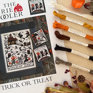 May include: A framed cross-stitch pattern with a Halloween theme, featuring a haunted house, witches, and the words "Trick or Treat." Several skeins of needlepoint silk thread in various colors, including cream, gold, orange, brown, black, and gray, are arranged alongside the pattern. Pumpkins and fall foliage complete the seasonal display.