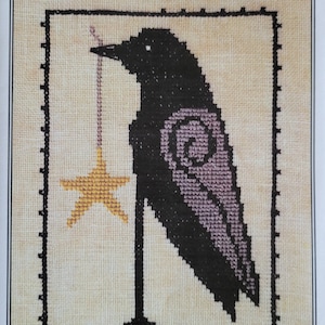 May include: Cross stitch pattern featuring a black crow holding a yellow star. The pattern is titled "Starring Russell Crow" and includes the website www.artful-offerings.com.