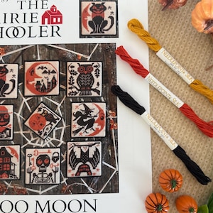 May include: A collection of Halloween-themed cross-stitch patterns featuring spooky cats, owls, and witches. The patterns are displayed on a printed page with the text "BOO MOON" and "The Prairie Schooler." Three skeins of needlepoint silk thread in black, red, and yellow are also visible.