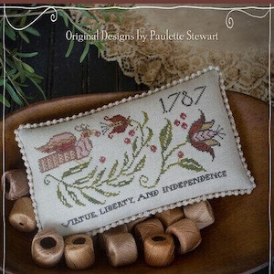 May include: A cross-stitched sampler with the text "1787" and "Virtue, Liberty, and Independence." The design includes a bird, flowers, and foliage in red, green, and pink hues. The sampler is displayed in a wooden bowl with spools of thread.