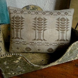 May include: A rectangular beige fabric pincushion featuring a brown cross-stitched pattern of three stylized trees and geometric borders, resting inside a weathered, distressed green metal container with a beaded rim and a curved front edge.