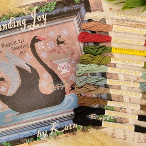 May include: A cross stitch pattern titled "The Sounding Joy" featuring a black swan with a blue and white wing, surrounded by snowflakes and reindeer. The pattern includes the text "Repeat the Sounding Joy" and the year "1810". The image also shows a variety of embroidery floss spools in different colors.