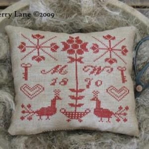 May include: A rectangular, beige pillow with red cross-stitch embroidery. The design includes floral and geometric patterns, keys, hearts, and birds. The text "M 18 70" is also visible. The pillow is placed in a wooden bowl with scissors.