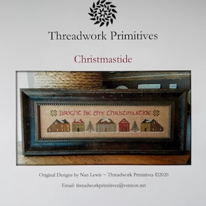 May include: A rectangular framed cross-stitch embroidery featuring a row of small houses and evergreen trees on a tan fabric background. The text "Bright Be thy Christmastide" is stitched in red above the houses. The frame is dark brown wood.