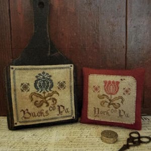 May include: A dark wooden cutting board with a square linen panel featuring a blue floral cross-stitch design and the text Bucks Co Pa, alongside a red fabric pincushion with a red floral cross-stitch design and the text York Co Pa, plus scissors.