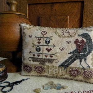 May include: A rectangular beige fabric pillow featuring a cross-stitched design of a black crow holding a red heart, a tiered stand with arrows and hearts, the number 14, and the text 'feb'. The pillow has a textured brown border trim.