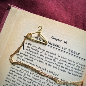 May include: A gold-colored metal hanger-shaped bookmark rests on an open book. The book's page displays the title "SEXUAL FREEING OF WOMAN" and text. A delicate gold chain is draped across the page, adding a decorative touch to the literary theme.