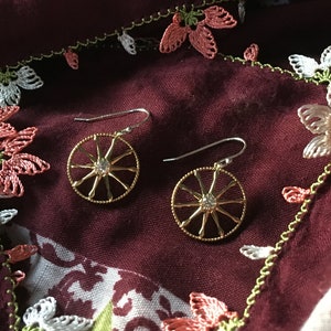 May include: A pair of gold earrings with a circular design featuring a sparkling stone in the center. The earrings have a delicate, intricate design and are suspended from silver hooks.