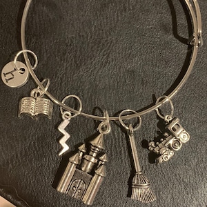Puede incluir: Un brazalete de plata con dijes que incluyen un libro, un rayo, un castillo, una escoba y un tren. El brazalete tiene un dije con la letra "H" en él.