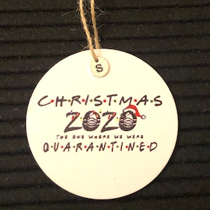 以下が含まれることがあります： 白い丸いクリスマスオーナメントに「CHRISTMAS 2020 THE ONE WHERE WE WERE QUARANTINED」の文字と、赤いサンタ帽子のついたフェイスマスクが描かれています。