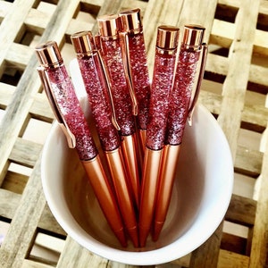 Op de afbeelding: Een set van zes roze gouden glitterpennen in een witte keramische beker. De pennen hebben een transparante cilinder gevuld met roze glitter.