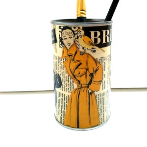 Puede incluir: Un bote de metal de estilo vintage con una ilustración de una mujer y un pájaro, reutilizado como porta lápices. Tiene un lápiz negro y un pincel dentro.