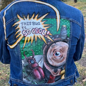 May include: A blue denim jacket with a hand-painted design on the back. The artwork features a large bug, an eye, and the text "THIS BUG IS.... SO COOL!" in a yellow and orange burst.