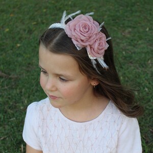 Op de afbeelding: Een wit hoofdband met twee roze stoffen rozen en witte veren. Het hoofdband wordt gedragen door een jong meisje met lang bruin haar.