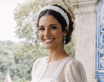 May include: A smiling person wearing a white wedding dress with sheer sleeves and lace cuffs. The dress features a high neckline and button details. A beaded headband and drop earrings complete the bridal look. The background includes a stone balcony and a blue and white tiled wall.