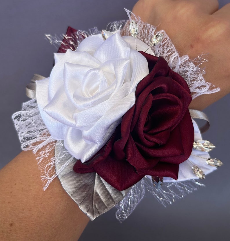 May include: A white and burgundy satin rose wrist corsage with silver and clear rhinestones and lace accents.