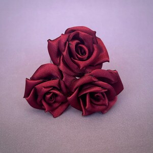 May include: Three burgundy satin roses arranged in a cluster. The roses are made of fabric and have a realistic appearance.