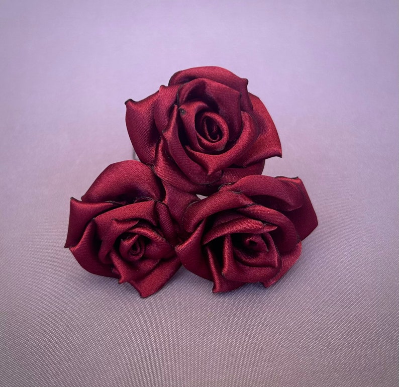 May include: Three burgundy satin roses arranged in a cluster. The roses are made of fabric and have a realistic appearance.