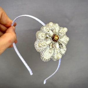 Puede incluir: Una diadema blanca con un gran adorno en forma de flor blanca. La flor est&aacute; hecha de perlas, pedrer&iacute;a y cuentas.