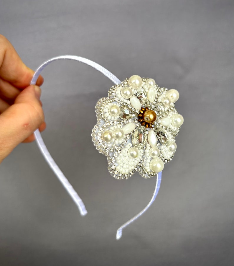 Puede incluir: Una diadema blanca con un gran adorno en forma de flor blanca. La flor est&aacute; hecha de perlas, pedrer&iacute;a y cuentas.