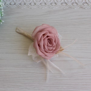 Op de afbeelding: Een roze stoffen roos met witte veren en een beige steel. De roos is een bruiloftscorsage.