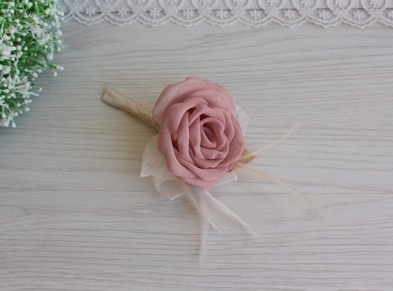 Op de afbeelding: Een roze stoffen roos met witte veren en een beige steel. De roos is een bruiloftscorsage.