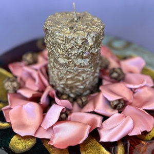 May include: A gold candle with a textured surface sits on a bed of pink satin rose petals. The petals are arranged in a circle around the candle, creating a romantic and elegant centerpiece.