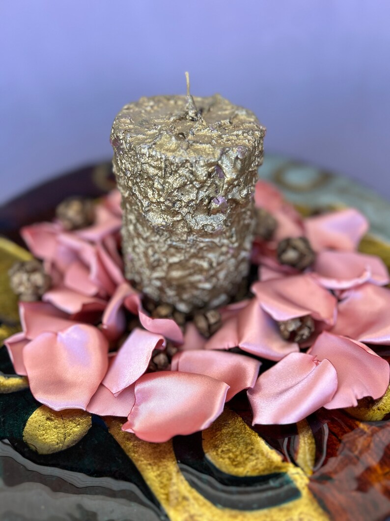 May include: A gold candle with a textured surface sits on a bed of pink satin rose petals. The petals are arranged in a circle around the candle, creating a romantic and elegant centerpiece.