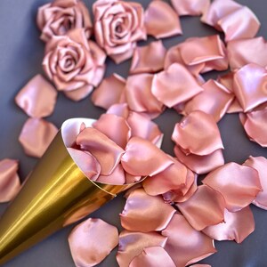 May include: A gold cone filled with rose petals, surrounded by loose rose petals on a gray surface. The petals are a soft blush pink color and made of satin.