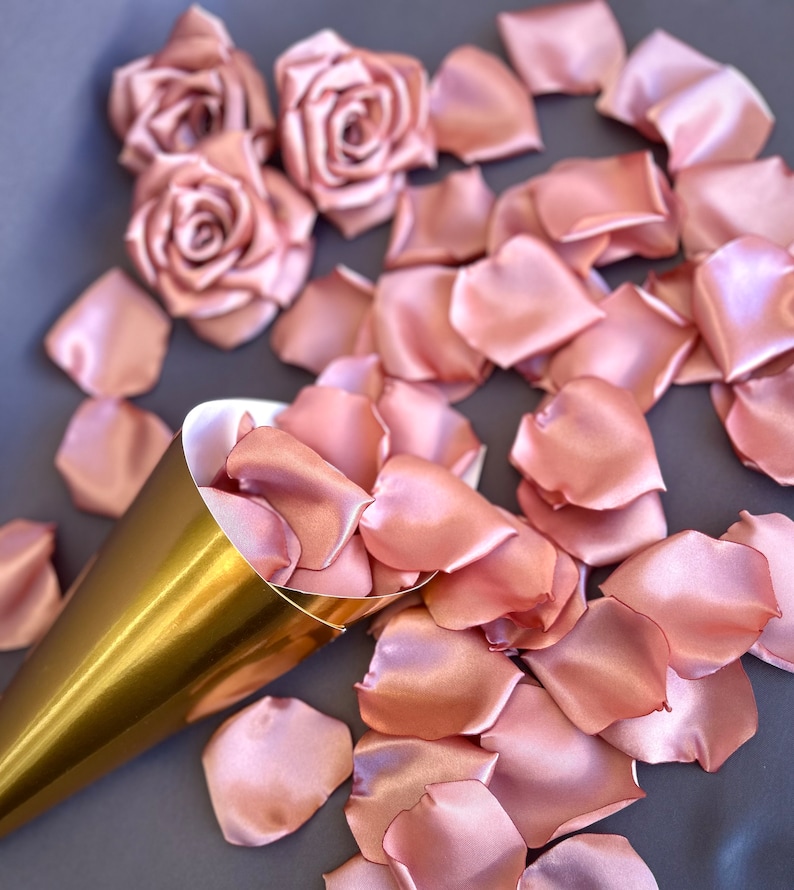 May include: A gold cone filled with rose petals, surrounded by loose rose petals on a gray surface. The petals are a soft blush pink color and made of satin.