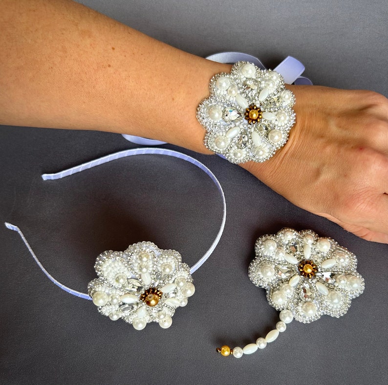Puede incluir: Un conjunto de tres accesorios para el cabello con forma de flor de cuentas blancas. El conjunto incluye una diadema, un brazalete y un broche. Las flores est&aacute;n hechas con perlas blancas y pedrer&iacute;a, y tienen un centro dorado.