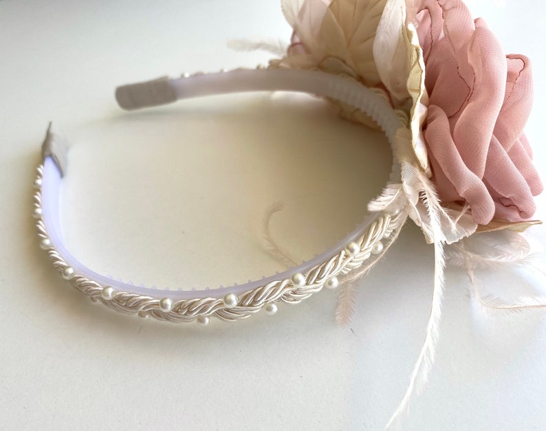 Op de afbeelding: Een wit hoofdband met een parelrand en een roze en cr&egrave;mekleurige bloemenversiering met veren.
