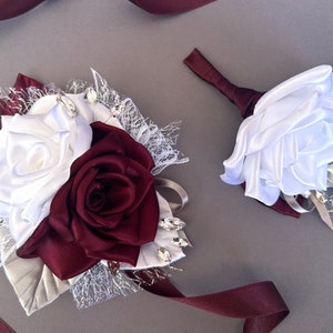 May include: A white and burgundy satin rose boutonniere and wrist corsage with lace and rhinestones.