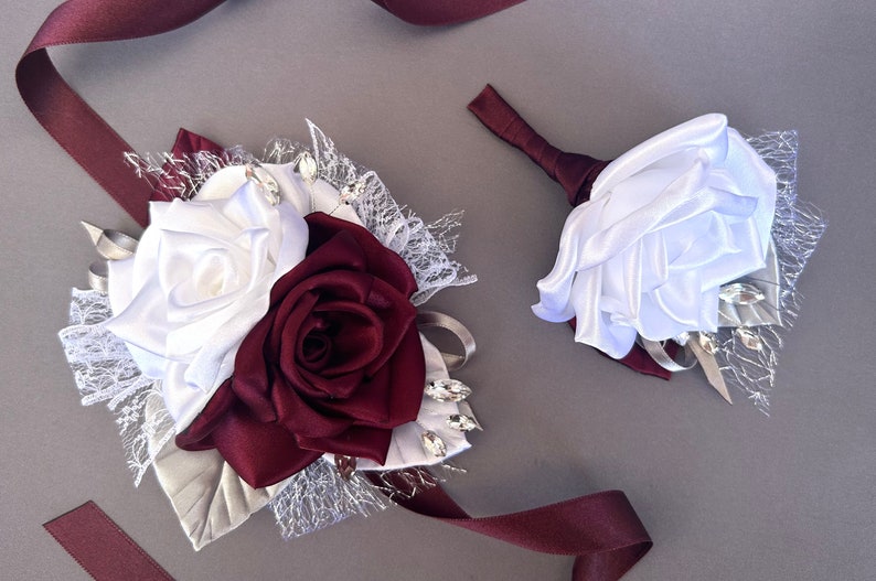 May include: A white and burgundy satin rose boutonniere and wrist corsage with lace and rhinestones.