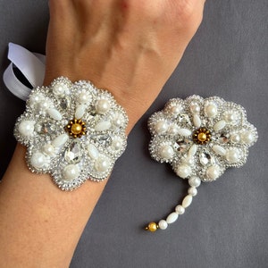 Puede incluir: Un par de broches blancas en forma de flor con adornos de perlas y cristales. La broche m&aacute;s grande se lleva en la mu&ntilde;eca, mientras que la m&aacute;s peque&ntilde;a tiene un tallo largo de perlas y cristales.