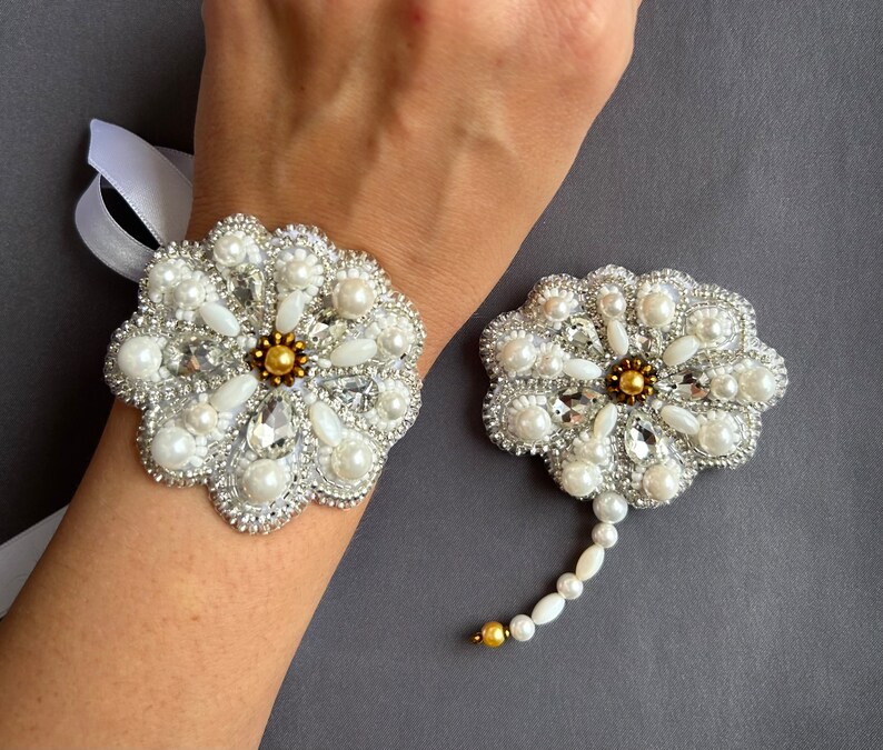 Puede incluir: Un par de broches blancas en forma de flor con adornos de perlas y cristales. La broche m&aacute;s grande se lleva en la mu&ntilde;eca, mientras que la m&aacute;s peque&ntilde;a tiene un tallo largo de perlas y cristales.
