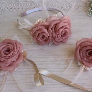 Op de afbeelding: Een set van drie roze stoffen bloemen haaraccessoires. De set bevat een hoofdband met twee bloemen, een polscorsage en een kleinere corsage. De bloemen zijn gemaakt van zacht stof en hebben een delicate, vrouwelijke uitstraling.