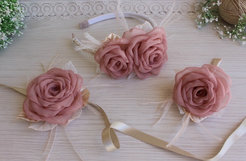 Op de afbeelding: Een set van drie roze stoffen bloemen haaraccessoires. De set bevat een hoofdband met twee bloemen, een polscorsage en een kleinere corsage. De bloemen zijn gemaakt van zacht stof en hebben een delicate, vrouwelijke uitstraling.