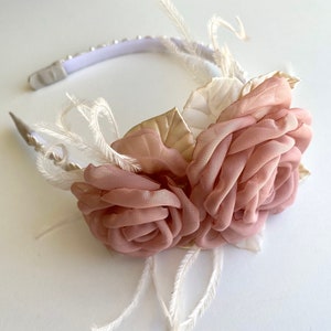 Op de afbeelding: Een roze bloemenhoofdband met witte veren en gouden accenten. De hoofdband is gemaakt van een wit elastisch band en heeft een delicaat, vrouwelijk ontwerp.