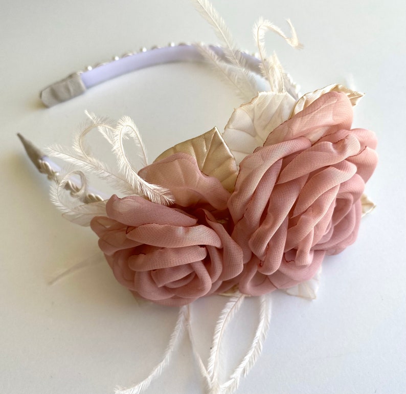 Op de afbeelding: Een roze bloemenhoofdband met witte veren en gouden accenten. De hoofdband is gemaakt van een wit elastisch band en heeft een delicaat, vrouwelijk ontwerp.