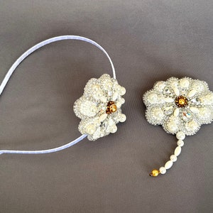 Puede incluir: Una diadema blanca con una flor de perlas y cristales. Un broche a juego de flores de perlas y cristales con una larga cola de perlas.